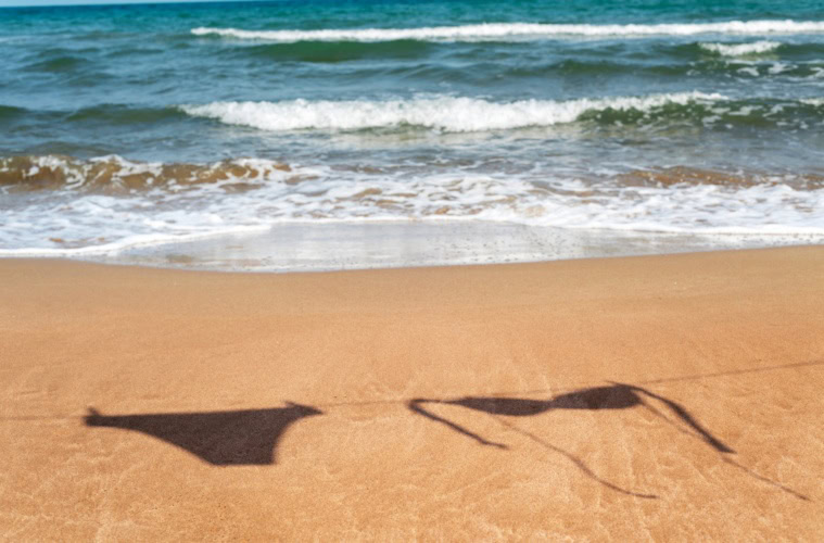 Auf dem Sand eines sommerlichen Strands ist der Schatten eines Bikinis an einer Wäscheleine zu sehen.