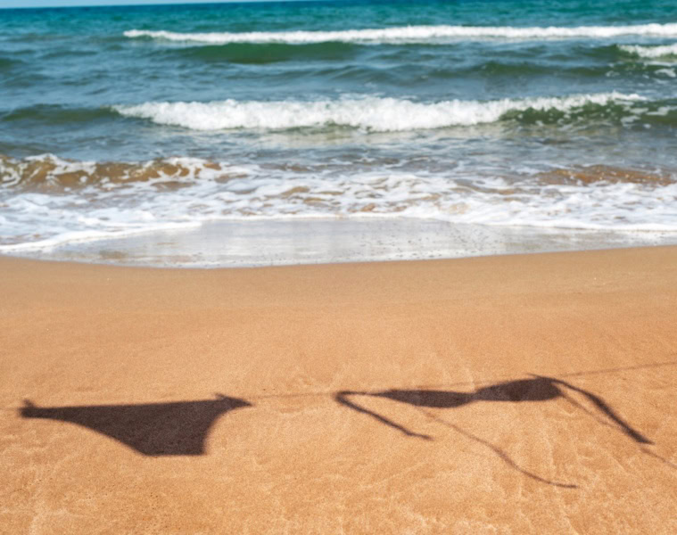 Auf dem Sand eines sommerlichen Strands ist der Schatten eines Bikinis an einer Wäscheleine zu sehen.