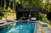 Ein moderner, in graue Betonplatten eingelassener Pool liegt in einem Wald. Am linken Rand zwei helle Liegen, vor Kopf eine hölzerne Sauna, vor der eine Frau in Bademantel und mit Tasche entlang läuft. Der Pool scheint inmitten eines Walds zu liegen.