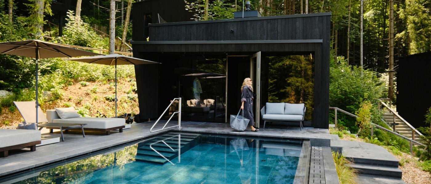 Ein moderner, in graue Betonplatten eingelassener Pool liegt in einem Wald. Am linken Rand zwei helle Liegen, vor Kopf eine hölzerne Sauna, vor der eine Frau in Bademantel und mit Tasche entlang läuft. Der Pool scheint inmitten eines Walds zu liegen.