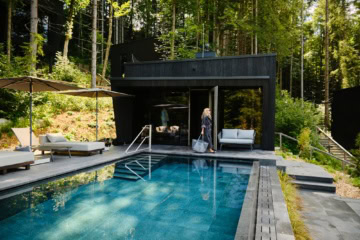 Ein moderner, in graue Betonplatten eingelassener Pool liegt in einem Wald. Am linken Rand zwei helle Liegen, vor Kopf eine hölzerne Sauna, vor der eine Frau in Bademantel und mit Tasche entlang läuft. Der Pool scheint inmitten eines Walds zu liegen.