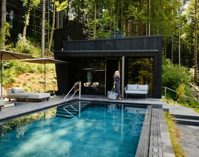 Ein moderner, in graue Betonplatten eingelassener Pool liegt in einem Wald. Am linken Rand zwei helle Liegen, vor Kopf eine hölzerne Sauna, vor der eine Frau in Bademantel und mit Tasche entlang läuft. Der Pool scheint inmitten eines Walds zu liegen.