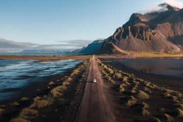 Drohnenaufnahme eines weißen Autos, das über eine langezogene Schotterpiste auf Island Richtung Horizont fährt, wo sich zur rechten ein gewaltiges Gebirge auftürmt.