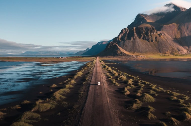 Drohnenaufnahme eines weißen Autos, das über eine langezogene Schotterpiste auf Island Richtung Horizont fährt, wo sich zur rechten ein gewaltiges Gebirge auftürmt.