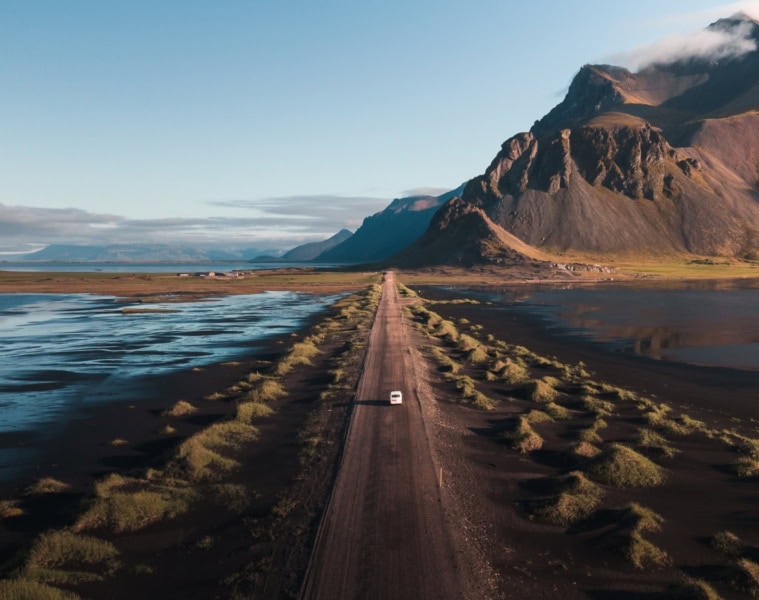 Drohnenaufnahme eines weißen Autos, das über eine langezogene Schotterpiste auf Island Richtung Horizont fährt, wo sich zur rechten ein gewaltiges Gebirge auftürmt.