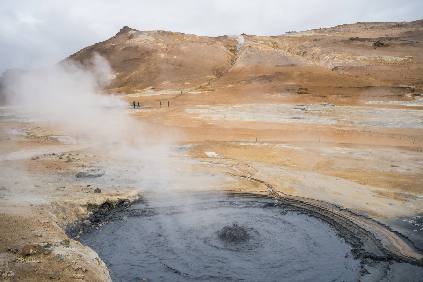 Inmitten einer mondartigen Gebirgslandschaft liegt ein kreisrunder, mit Wasser gefüllter Krater, aus dessen Mitte blubbernd eine Dampfwolke aussteigt.