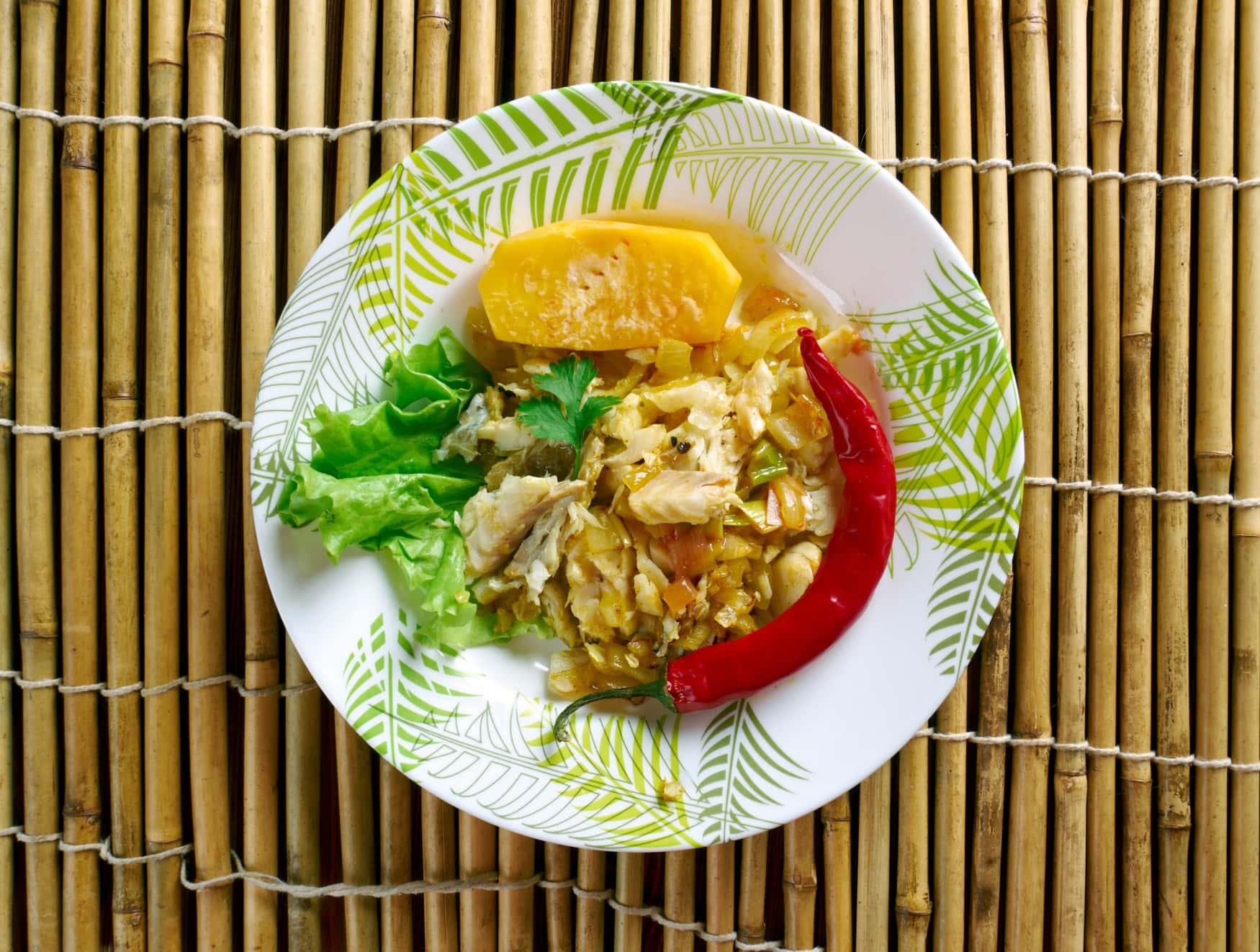 Auf einem Teller ist das jamaikanische Nationalgericht "Ackee and Saltfish" angerichtet, mit einer Kartoffel und einer Chilli garniert. Der Teller steht auf einer Bast-Unterlage.
