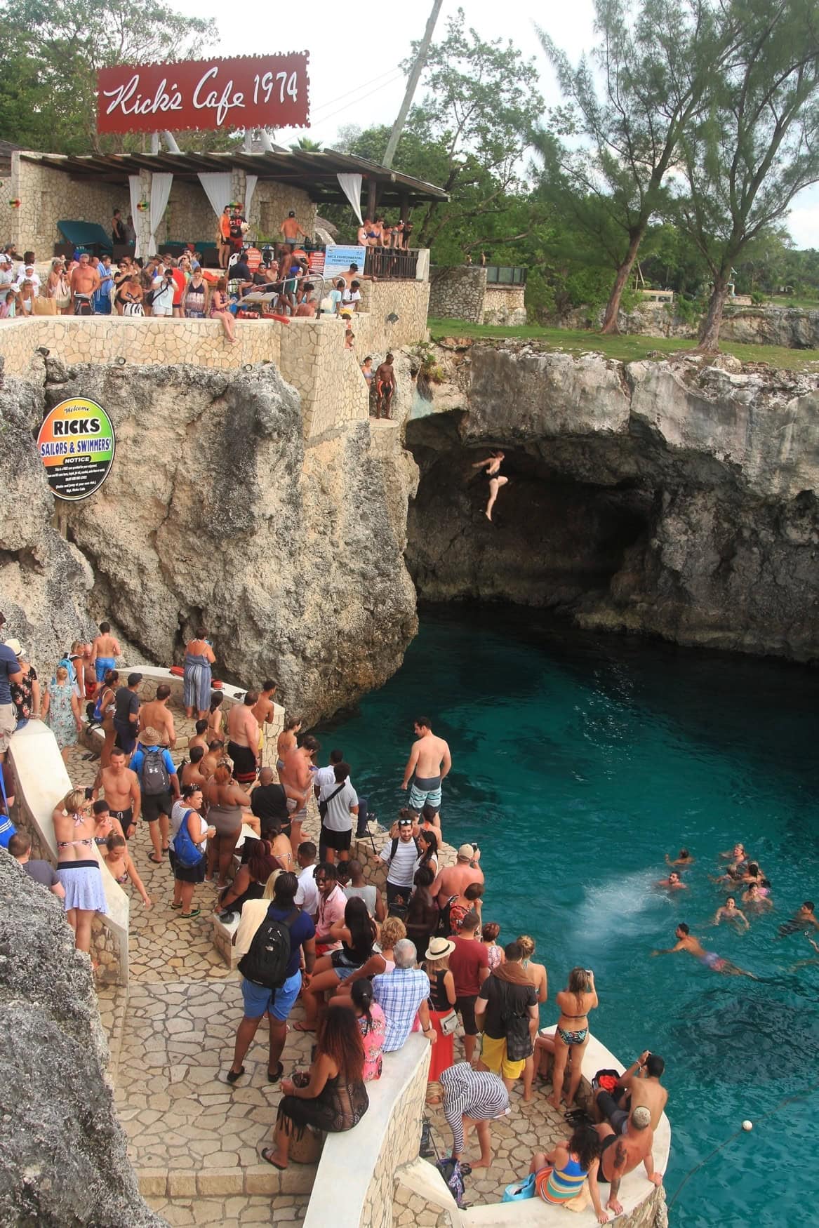 Um eine halbkreisförmige Klippe stehen viele Menschen gedrängt, meist in Schwimmkleidung gekleidet. Eine Frau ist gerade gesprungen und befindet sich im Flug ins Wasser. Links im Hintergrund ist ein Café mit dem Schild "Rick's Cafe 1974" sichtbar.
