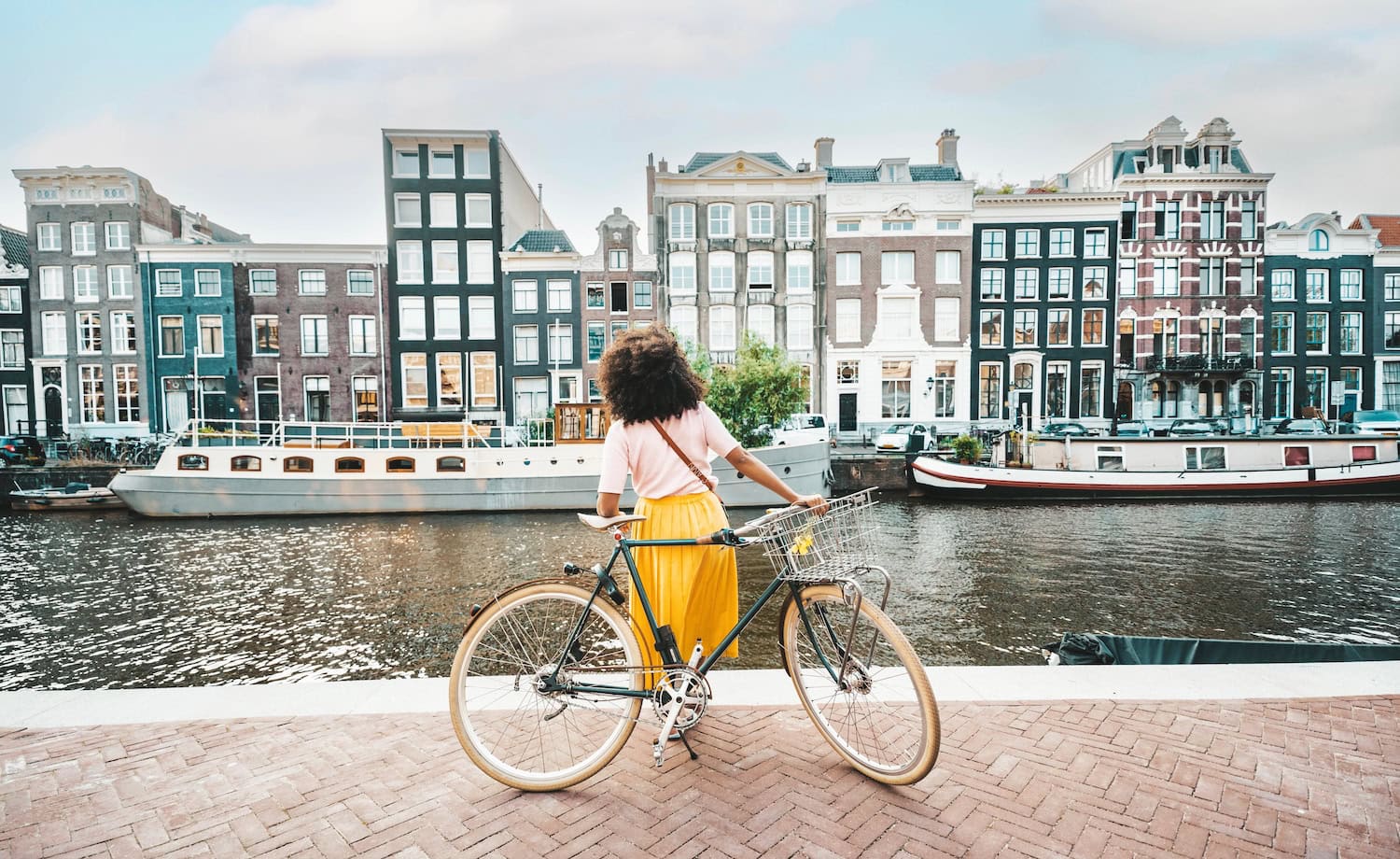 Junge Frau im gelben Rock und weißen Shirt lehnt sich an ihr Fahrrad und schaut auf die Grachtenhäuser in Amsterdam.