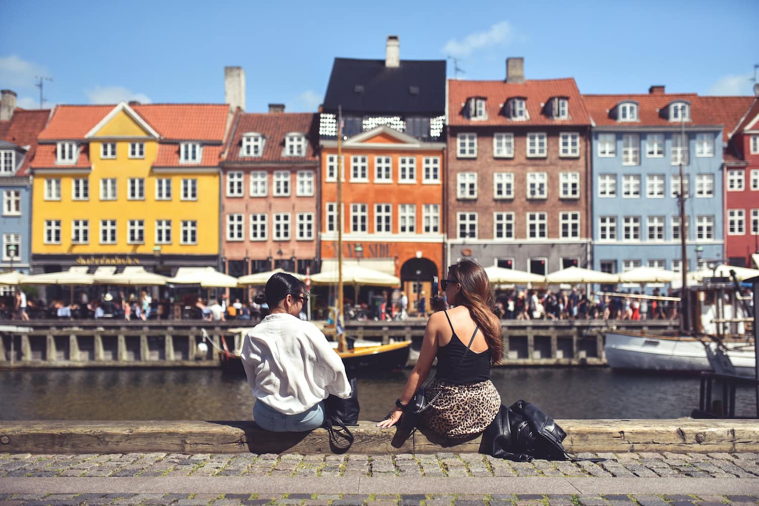 Zwei Frauen sitzen am Hafen in Kopenhagen und blicken auf Wasser und Häuser in Nyhavn.