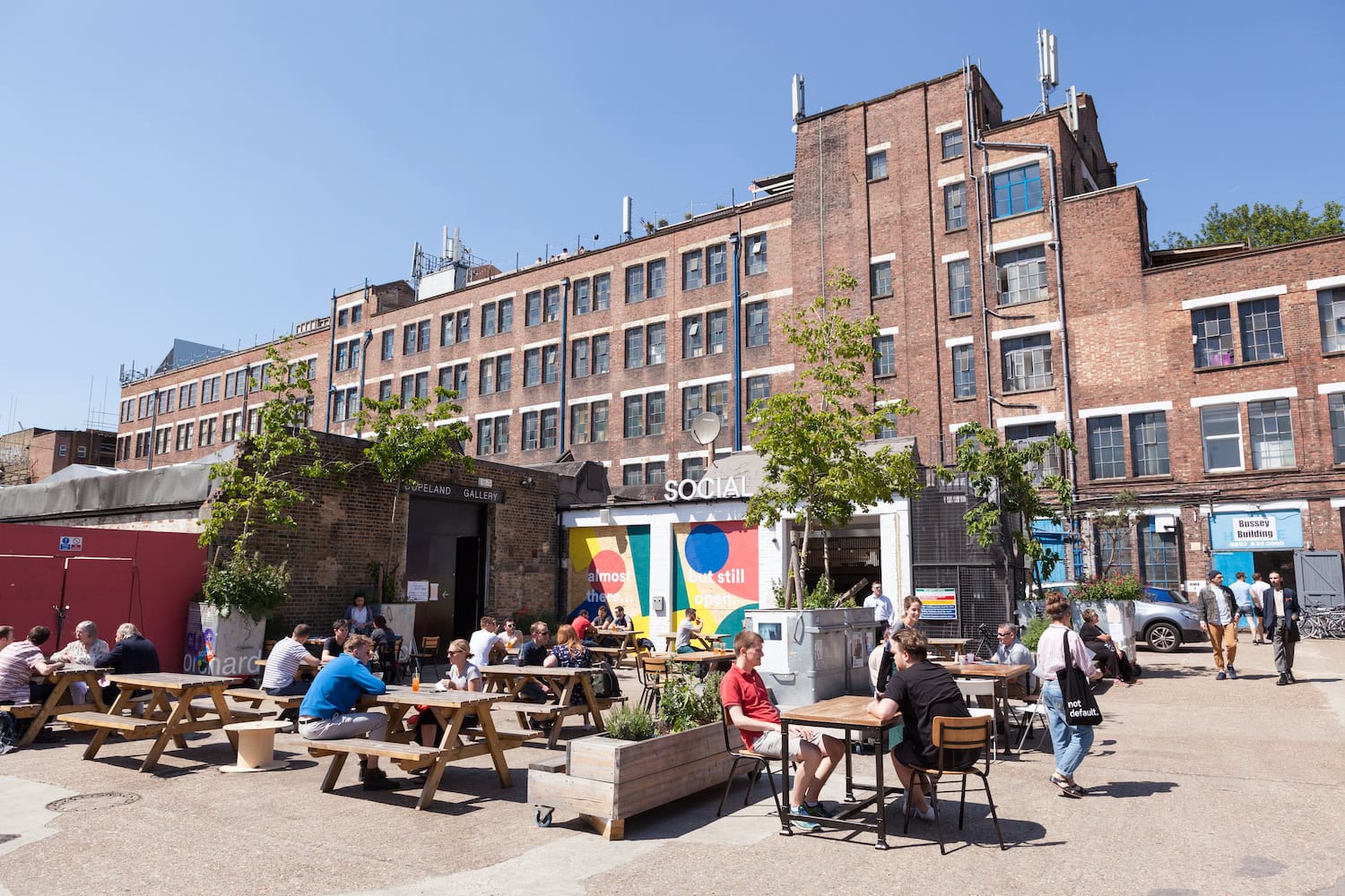 London Peckham: Menschen sitzen in der Sonne an Bänken draußen vor einem großen fabrikartigem Gebäude.
