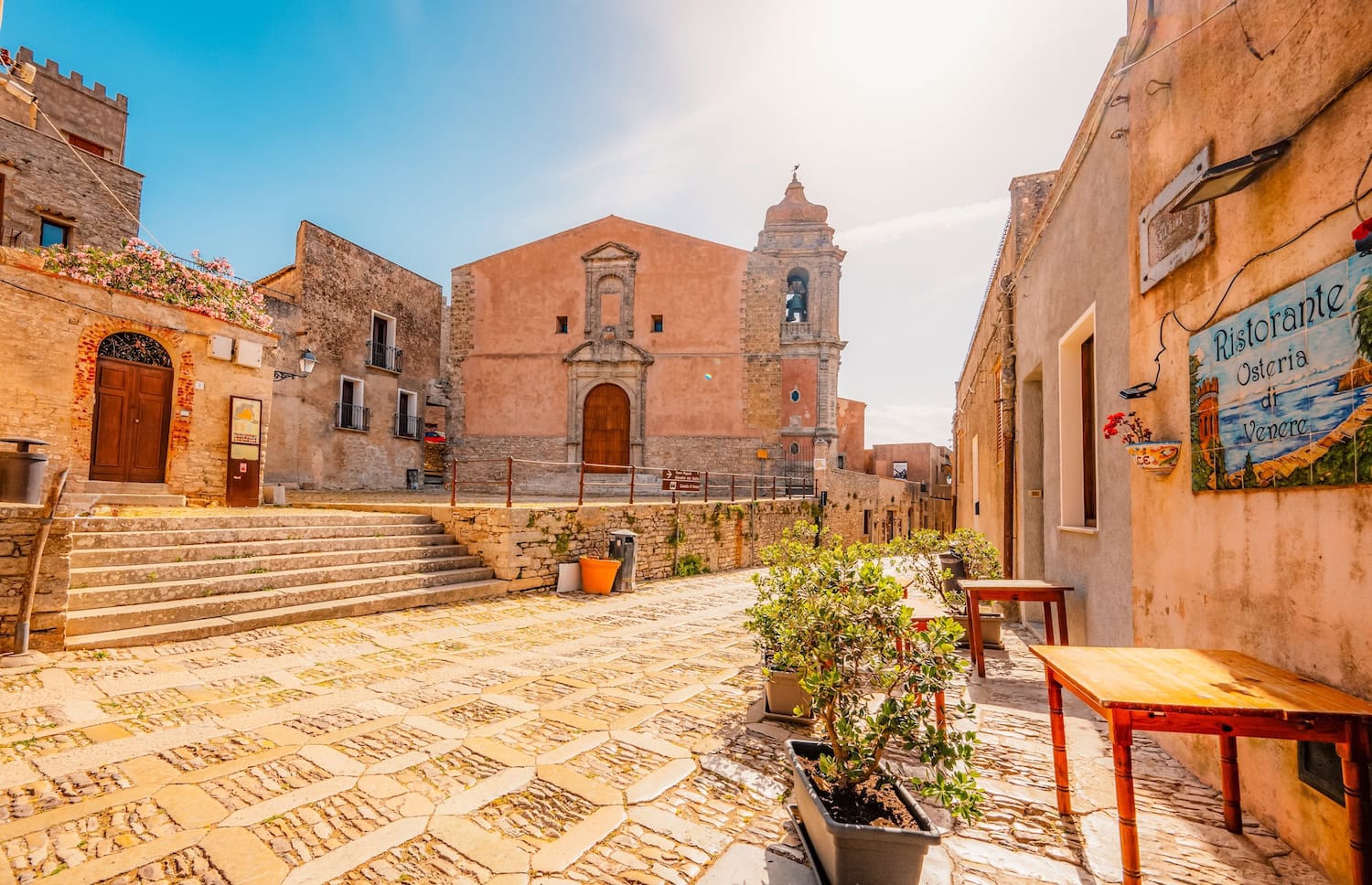 Platz im Örtchen Trapani in Sizilien im Sommer. Der Platz ist menschenleer, im Vordergrund eine alte Kirche, umgeben von alten Häusern. Links ist ein Schild zu einer Osteria mit Tischen davor.