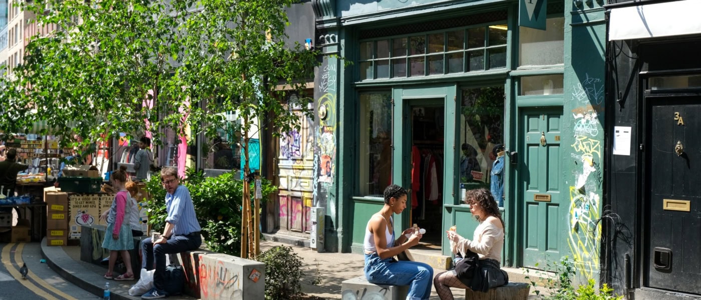Vor einem grün angestreichenen Café in Shoreditch in London sitzen zwei junge Frauen in der Sonne und essen ein Sandwich.