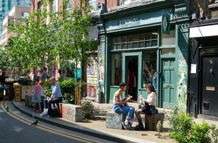Vor einem grün angestreichenen Café in Shoreditch in London sitzen zwei junge Frauen in der Sonne und essen ein Sandwich.