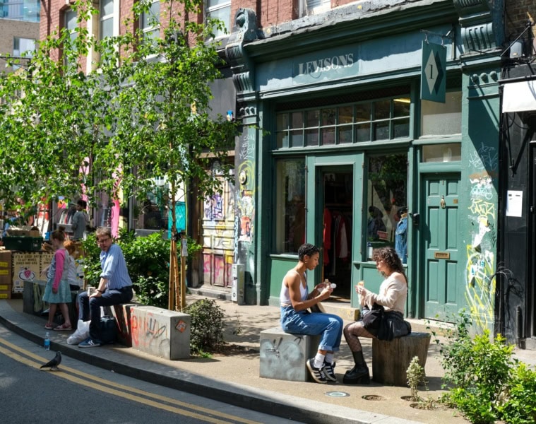 Vor einem grün angestreichenen Café in Shoreditch in London sitzen zwei junge Frauen in der Sonne und essen ein Sandwich.