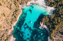 Senkrechte Drohnenaufnahme der Cala en Turqueta mit gestochen klarem türkisem Wasser, weißem Sandstrand, einem ankernden Segelboot und dichtem Pinienwald.