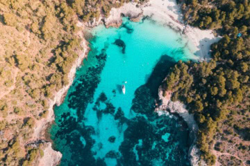 Senkrechte Drohnenaufnahme der Cala en Turqueta mit gestochen klarem türkisem Wasser, weißem Sandstrand, einem ankernden Segelboot und dichtem Pinienwald.