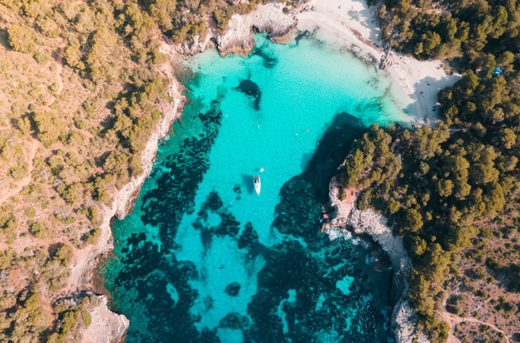 Senkrechte Drohnenaufnahme der Cala en Turqueta mit gestochen klarem türkisem Wasser, weißem Sandstrand, einem ankernden Segelboot und dichtem Pinienwald.
