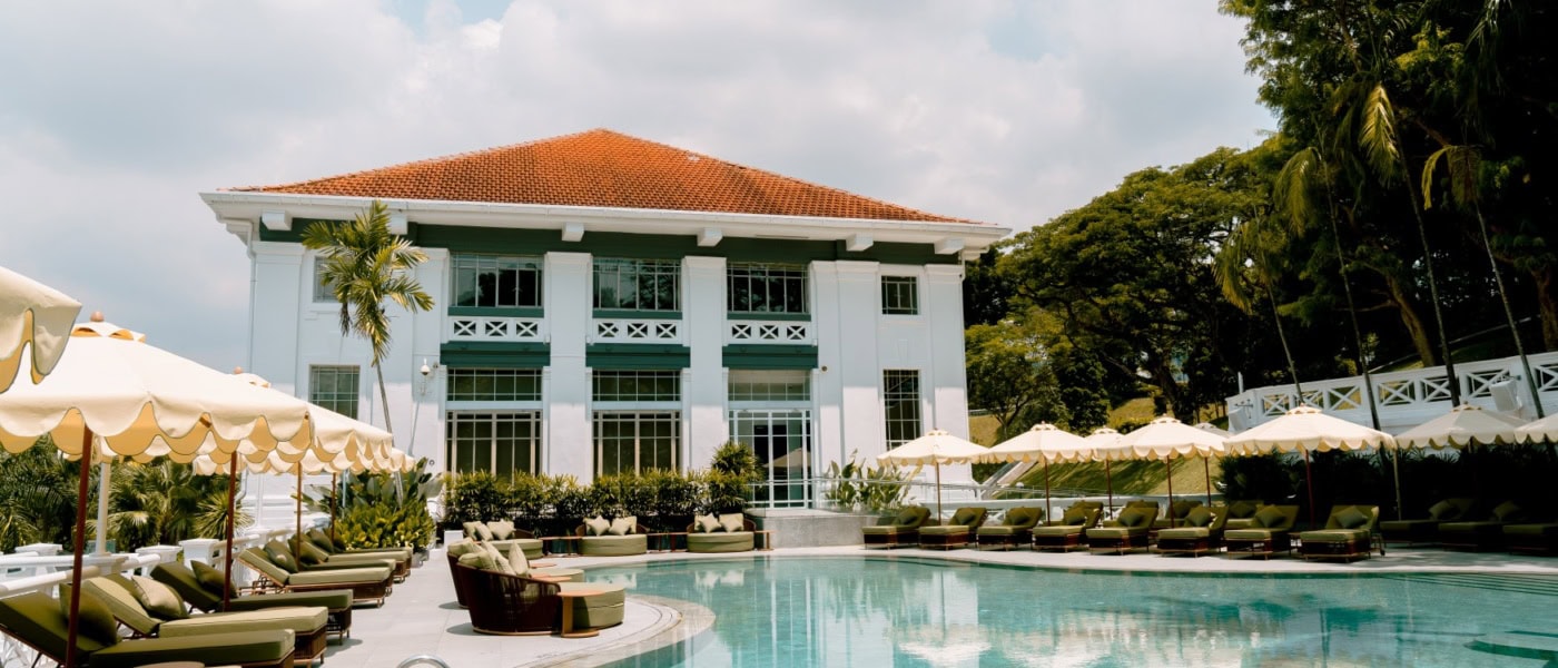 Vor einem Hotel in weißem Kolonialstil in grüner Umgebung liegt ein geschwungener Pool mit Liegen an beiden Seiten.