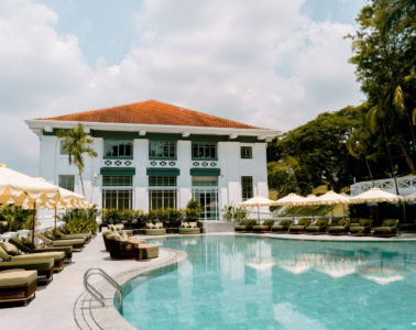 Vor einem Hotel in weißem Kolonialstil in grüner Umgebung liegt ein geschwungener Pool mit Liegen an beiden Seiten.