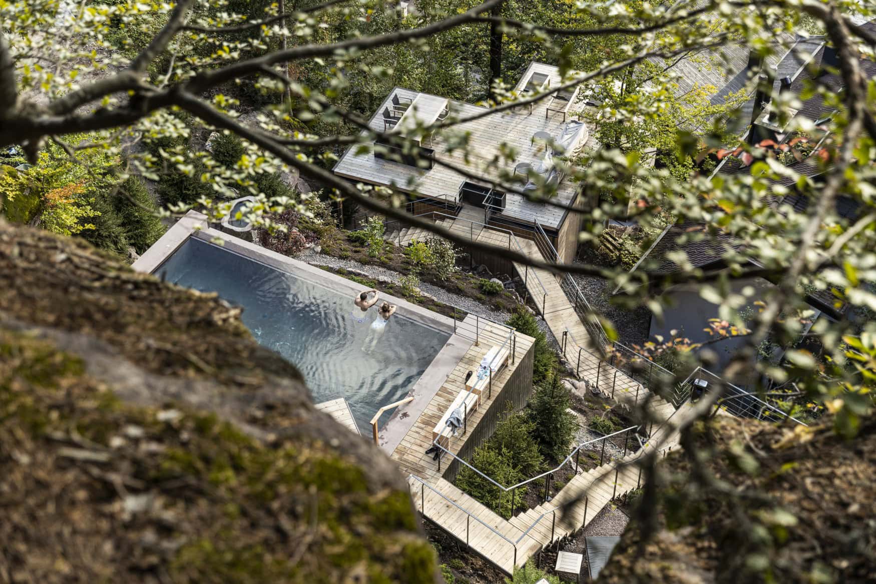 Blick von oben auf einen in einen Waldhang eingelassenen Onsen-Pool. Vom Pool führt eine Holztreppe den Hang hinab zu einer Terrasse mit Sitzgelegenheiten und Liegestühlen.