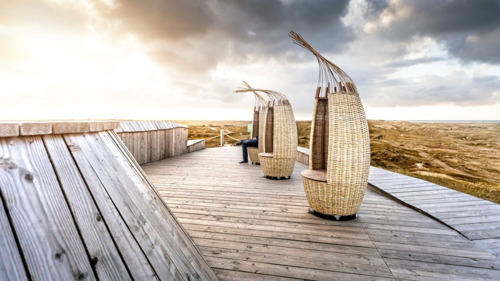 Auf einer hölzernen Plattform inmitten einiger Dünen stehen drei hohe, schmale Strandkörbe.