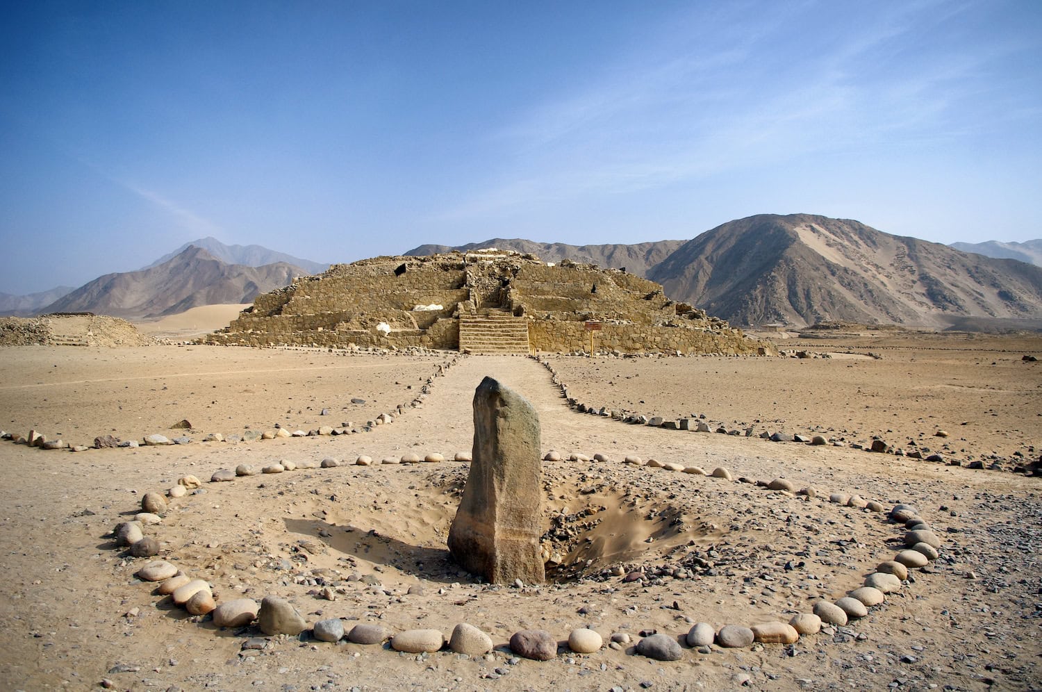 Die Archäologische Zone von Caral in Peru, Ruinen aus Stein inmitten einer Steinwüste, im Vordergrund ein hoher länglicher Stein mit einer runden Umrandung aus kleinen Steinen.