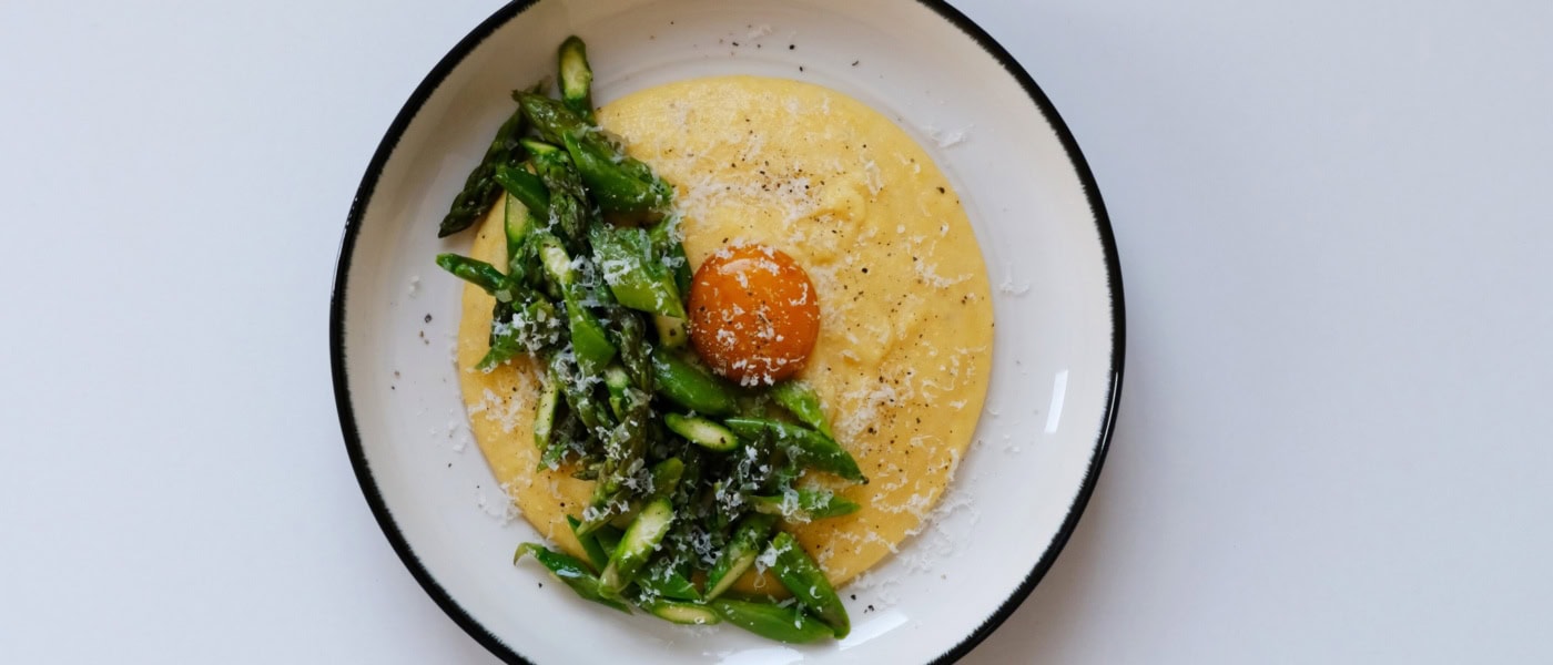In einem tiefen Teller ist eine Portion Polenta mit gehacktem, grünen Spargel angerichtet. Genau in der Mitte ist ein pochiertes Eigelb platziert, über alles wurde Käse gerieben.