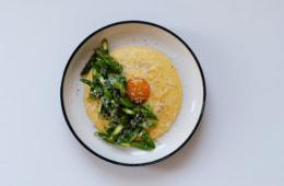 In einem tiefen Teller ist eine Portion Polenta mit gehacktem, grünen Spargel angerichtet. Genau in der Mitte ist ein pochiertes Eigelb platziert, über alles wurde Käse gerieben.