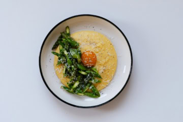 In einem tiefen Teller ist eine Portion Polenta mit gehacktem, grünen Spargel angerichtet. Genau in der Mitte ist ein pochiertes Eigelb platziert, über alles wurde Käse gerieben.