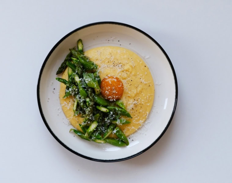 In einem tiefen Teller ist eine Portion Polenta mit gehacktem, grünen Spargel angerichtet. Genau in der Mitte ist ein pochiertes Eigelb platziert, über alles wurde Käse gerieben.