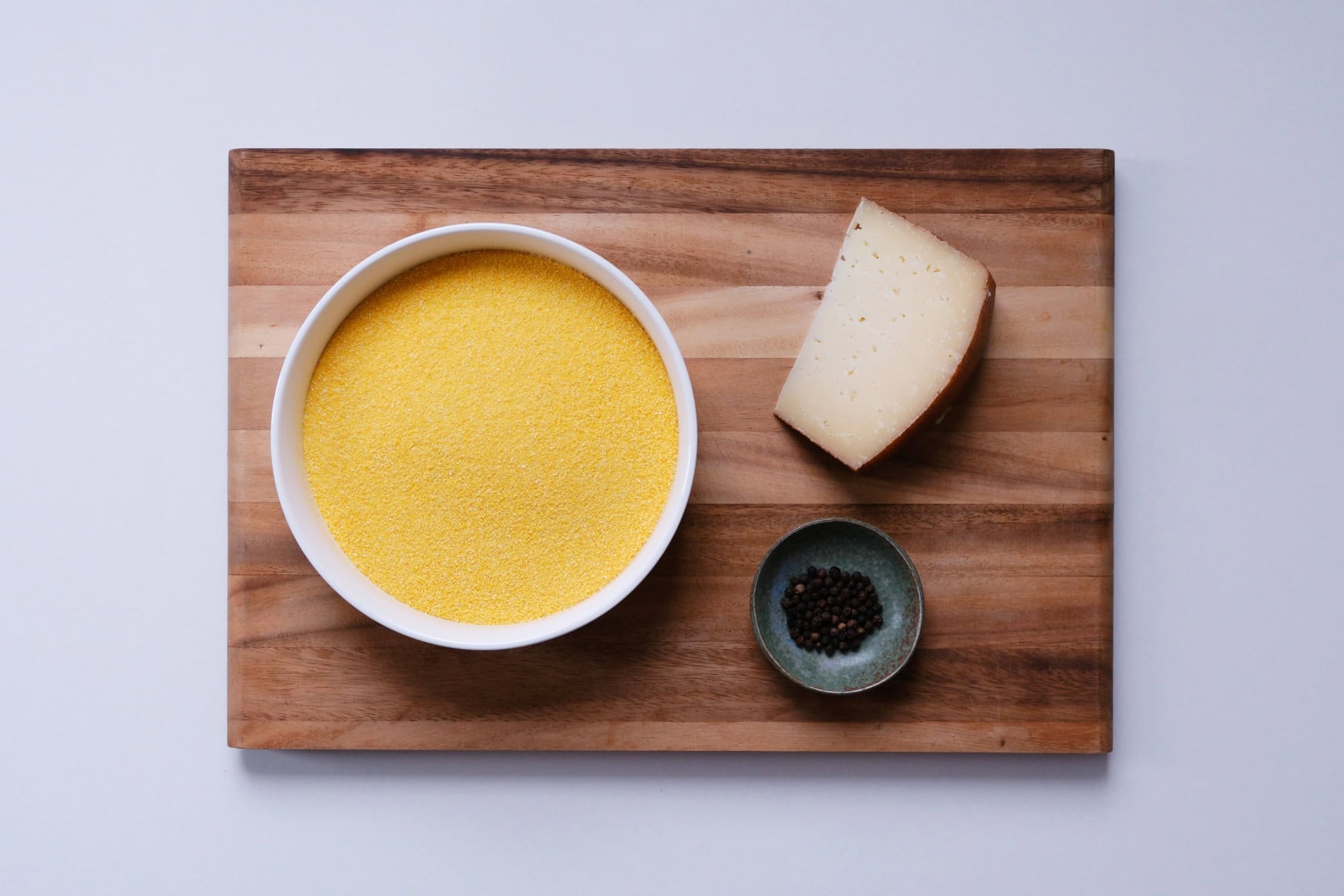 Auf einem Holzbrett liegen eine große Schüssel Polenta, eine kleine Schüssel mit schwarzem Pfeffer und ein Stück Peccorino.
