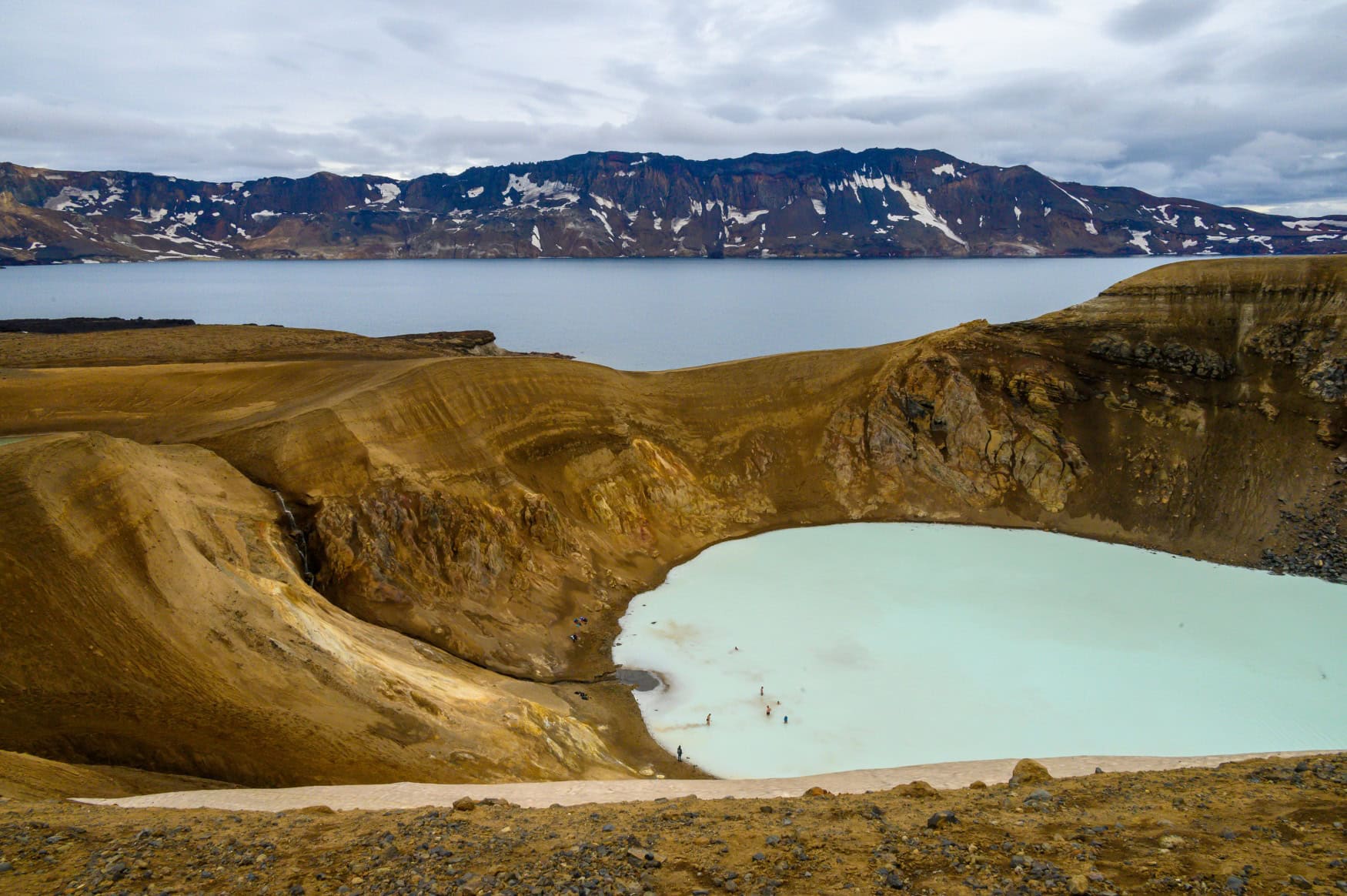 Ein Vulkankrater, gefüllt mit hellblauem Wasser, in dem einige Menschen schwimmen. Der Kraterrand trennt das Wasser im Krater von einem großen See, der dahinter liegt.