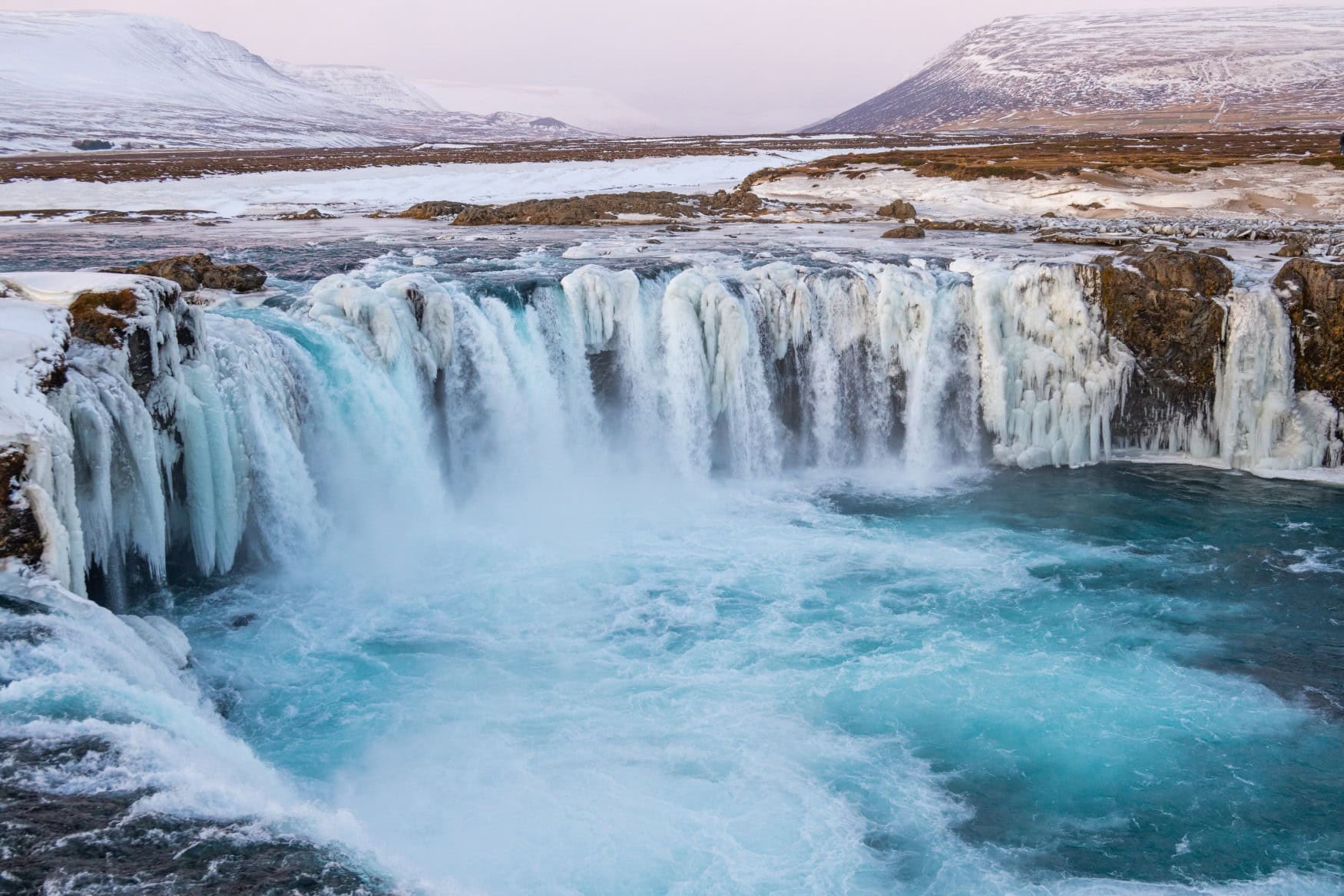 Ein Wasserfall in einer verschneiten isländischen Landschaft stürzt von einer halbrunden Klippe in die Tiefe.