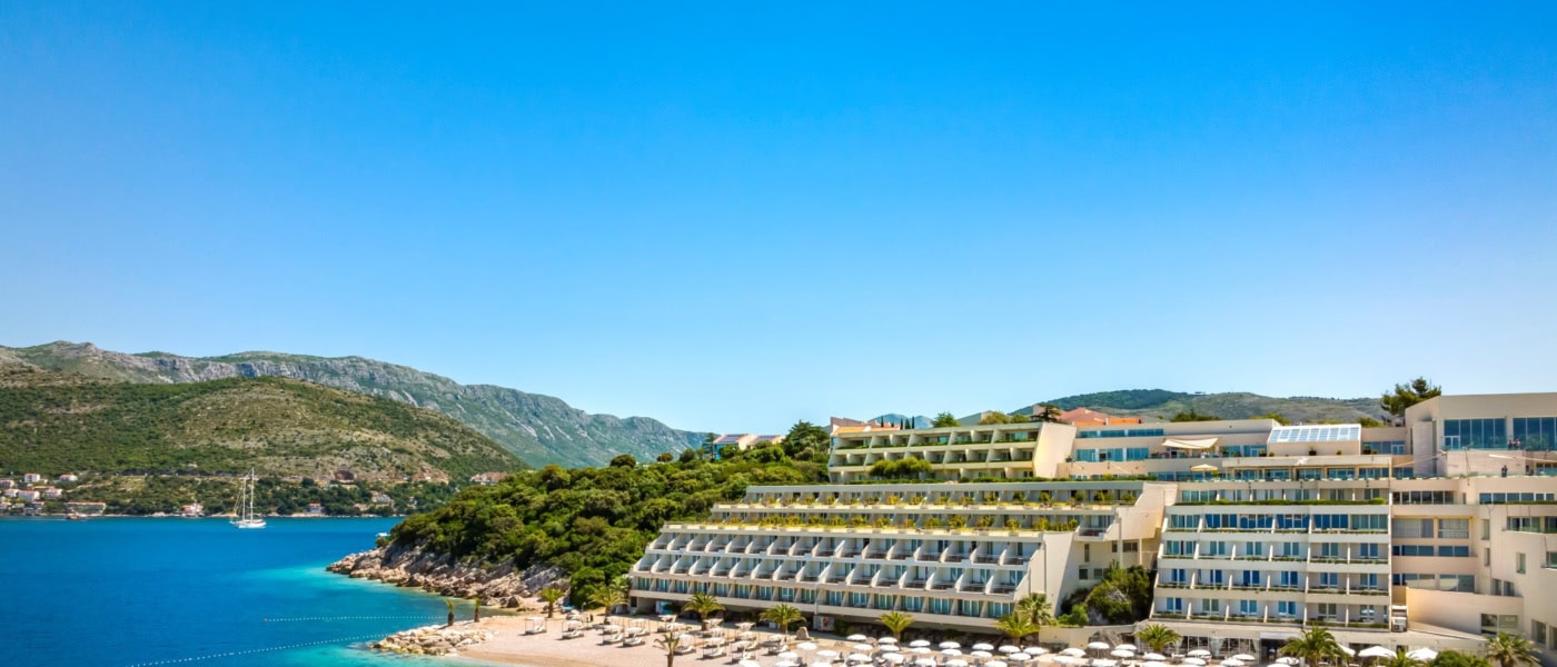 Ein Strand in Kroatien unter blauem Himmel. Zur rechten liegt ein großer, weißer Hotelkomplex im Licht der Mittagssonne.