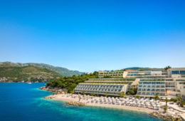Ein Strand in Kroatien unter blauem Himmel. Zur rechten liegt ein großer, weißer Hotelkomplex im Licht der Mittagssonne.