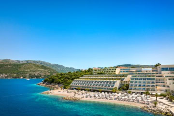 Ein Strand in Kroatien unter blauem Himmel. Zur rechten liegt ein großer, weißer Hotelkomplex im Licht der Mittagssonne.