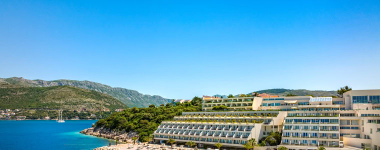 Ein Strand in Kroatien unter blauem Himmel. Zur rechten liegt ein großer, weißer Hotelkomplex im Licht der Mittagssonne.