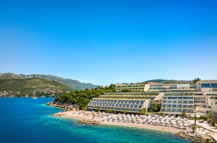 Ein Strand in Kroatien unter blauem Himmel. Zur rechten liegt ein großer, weißer Hotelkomplex im Licht der Mittagssonne.