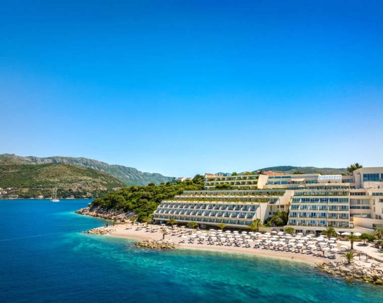 Ein Strand in Kroatien unter blauem Himmel. Zur rechten liegt ein großer, weißer Hotelkomplex im Licht der Mittagssonne.