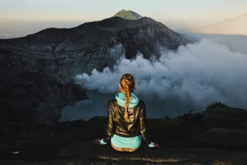 Vulkan-Trekking bei Sonnenaufgang: Junge Wanderin mit geflochtenem Haar sitzt am Kraterrand und blickt auf einen rauchenden Vulkan.