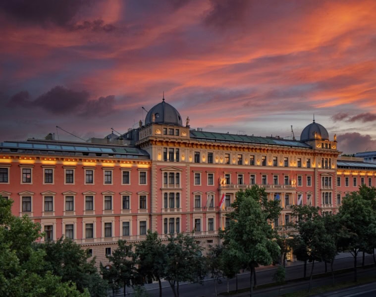 Das Anantara Palais Hansen Vienna Hotel von außen; ein klassizistischer Palas mit lachsroter Fassade, darüber der rote Abendhimmel.