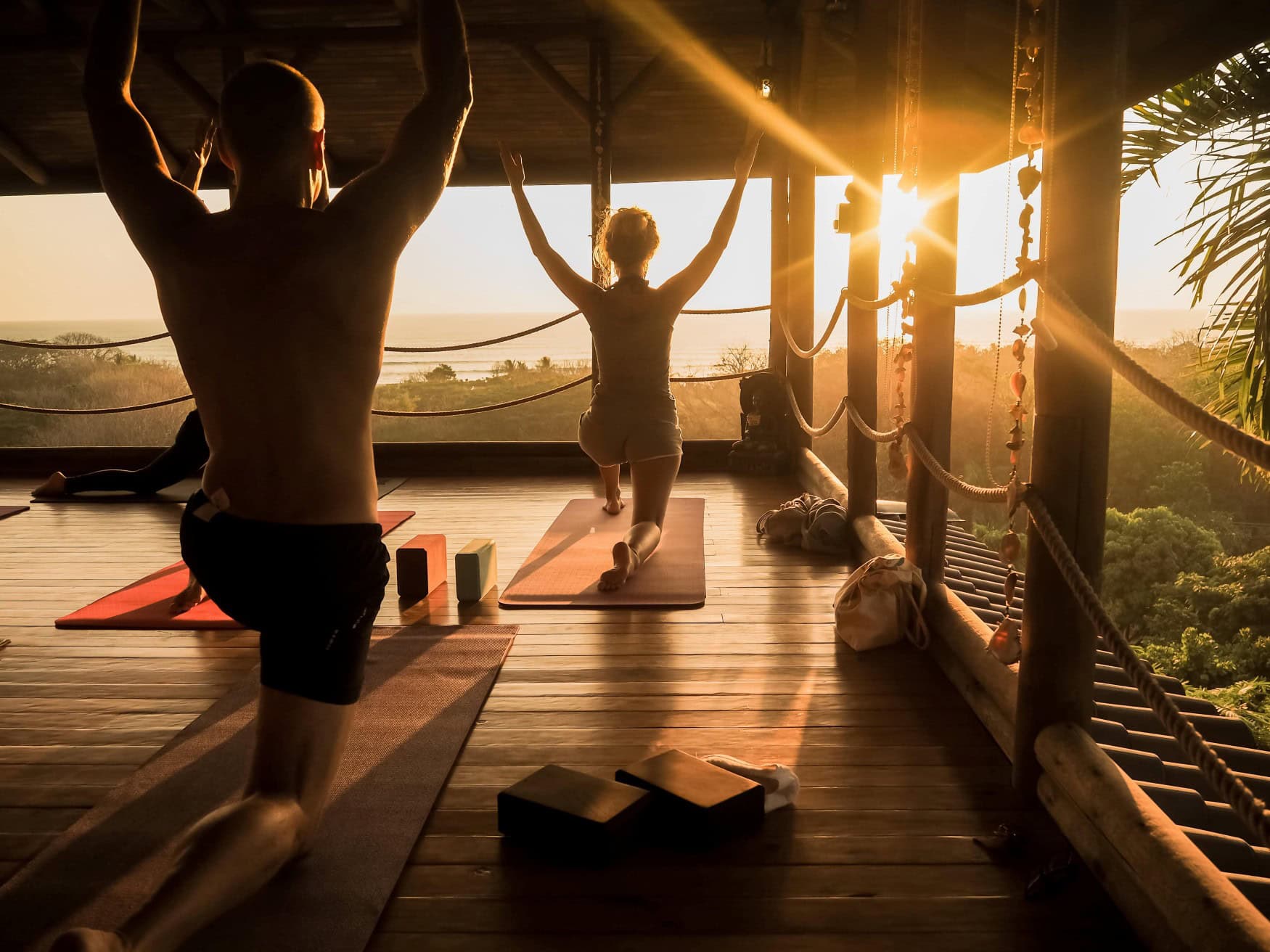 Yogagruppe von hinten beim Sonnenuntergang in einem offenen Gebäude aus Holz.