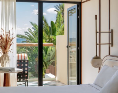 Bless Ibiza The Site - Premium Raum, im Vordergrund ein Bett mit glatten weißen Laken, links ein eleganter Beistelltisch mit Buch und Trockenblumen, die Balkontür ist geöffnet und zeigt das Meer, Palmen.