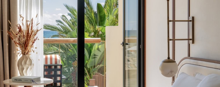 Bless Ibiza The Site - Premium Raum, im Vordergrund ein Bett mit glatten weißen Laken, links ein eleganter Beistelltisch mit Buch und Trockenblumen, die Balkontür ist geöffnet und zeigt das Meer, Palmen.