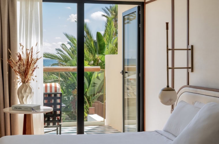 Bless Ibiza The Site - Premium Raum, im Vordergrund ein Bett mit glatten weißen Laken, links ein eleganter Beistelltisch mit Buch und Trockenblumen, die Balkontür ist geöffnet und zeigt das Meer, Palmen.