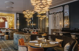 Elegantes Restaurant mit dunkelgrünen Wänden, opulenten goldenen Blüten-Kronleuchtern, gemusterten Polstersofas, samtbezogenen Sesseln und gedeckten Tischen auf orientalischem Teppich.