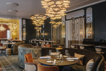Elegantes Restaurant mit dunkelgrünen Wänden, opulenten goldenen Blüten-Kronleuchtern, gemusterten Polstersofas, samtbezogenen Sesseln und gedeckten Tischen auf orientalischem Teppich.