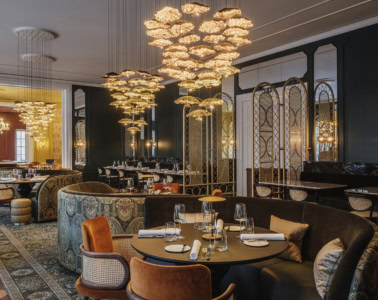 Elegantes Restaurant mit dunkelgrünen Wänden, opulenten goldenen Blüten-Kronleuchtern, gemusterten Polstersofas, samtbezogenen Sesseln und gedeckten Tischen auf orientalischem Teppich.