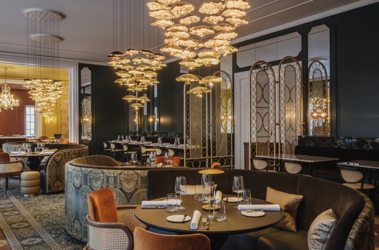 Elegantes Restaurant mit dunkelgrünen Wänden, opulenten goldenen Blüten-Kronleuchtern, gemusterten Polstersofas, samtbezogenen Sesseln und gedeckten Tischen auf orientalischem Teppich.
